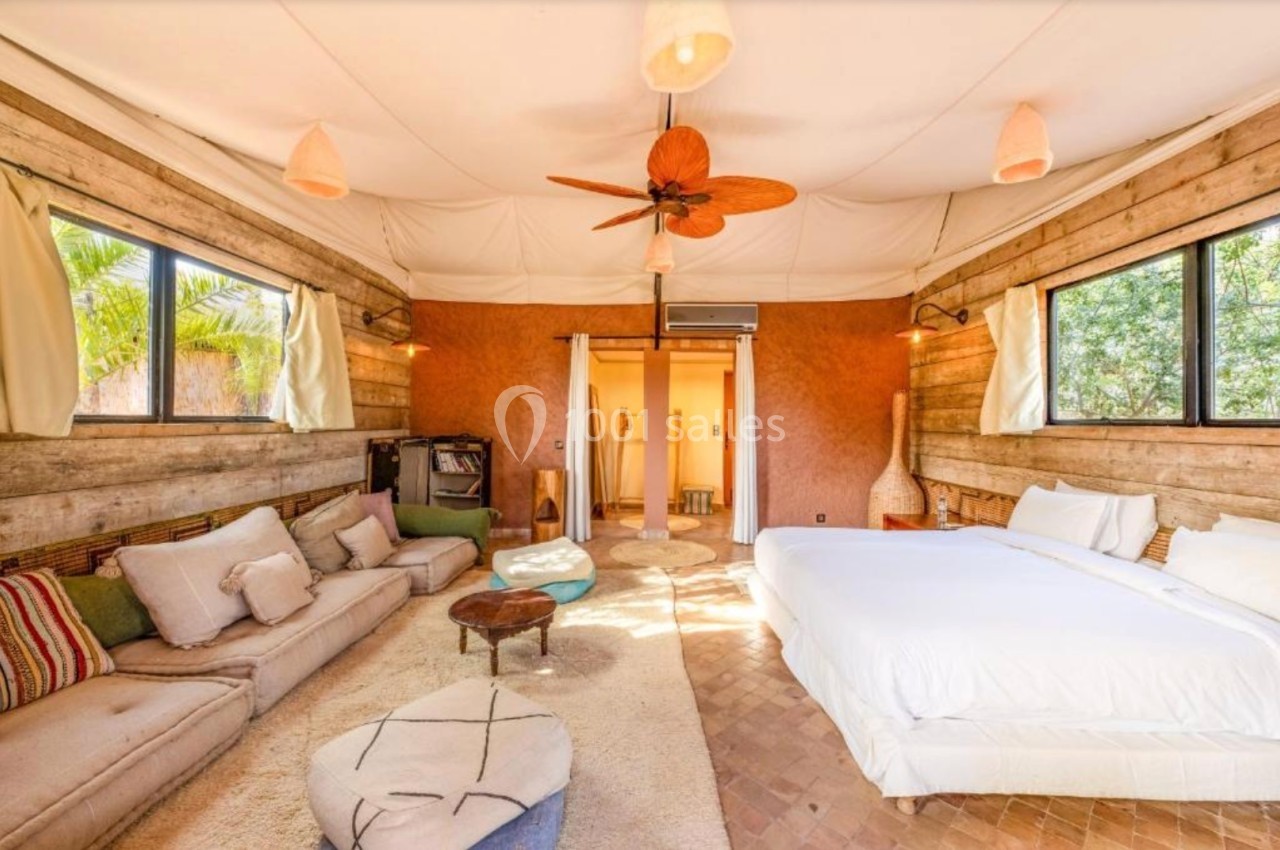 Chambre lumineuse avec lit double, canapé, tapis beige, ventilateur au plafond et grandes fenêtres donnant sur l'extérieur.