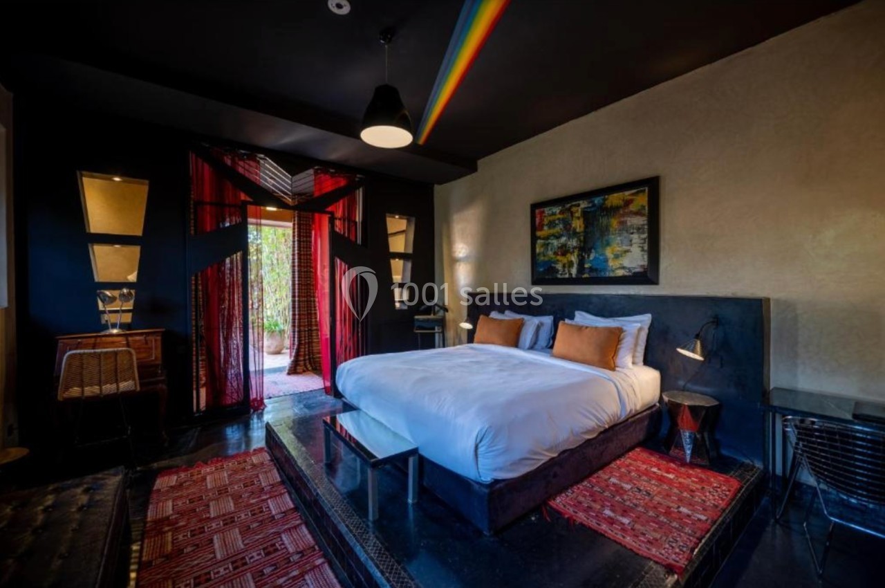 Chambre moderne avec lit double, décoration colorée, tapis rouges, et lumière naturelle entrant par une porte vitrée.