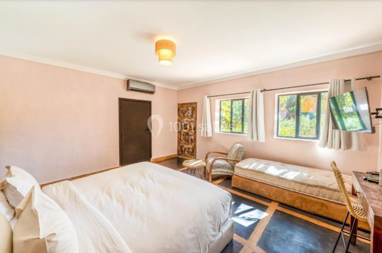 Chambre lumineuse avec lit double, méridienne, bureau, télévision murale et fenêtres donnant sur un jardin.