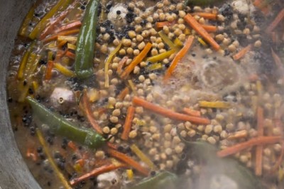 Un homme cuisine de grandes pièces de viande dans une marmite fumante en extérieur, portant un tablier et des gants.