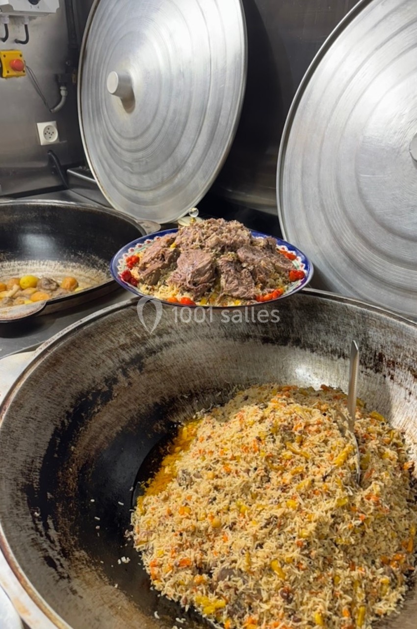 Plat de riz aux légumes et viande dans une grande marmite, avec un plat de viande et légumes en arrière-plan.