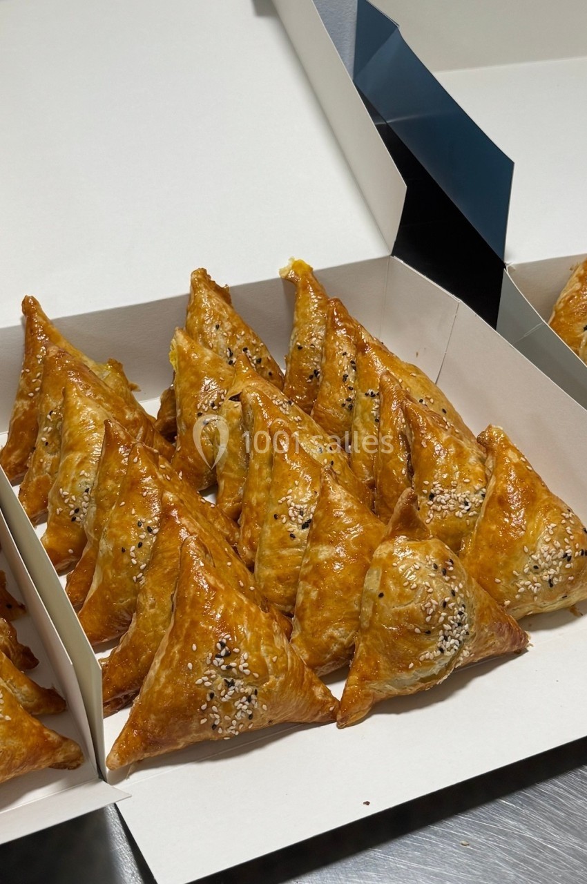 Assortiment de feuilletés dorés en forme triangulaire, garnis de graines de sésame, disposés dans une boîte ouverte.