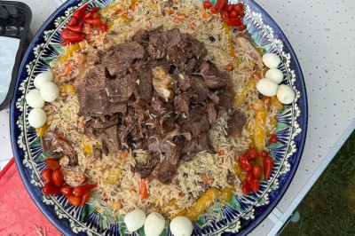 Un homme cuisine de grandes pièces de viande dans une marmite fumante en extérieur, portant un tablier et des gants.