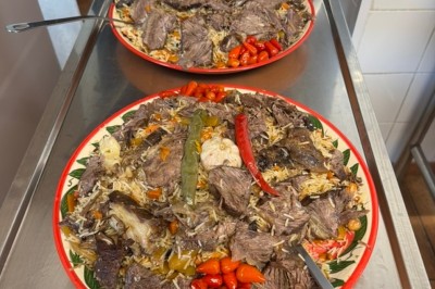 Un homme cuisine de grandes pièces de viande dans une marmite fumante en extérieur, portant un tablier et des gants.