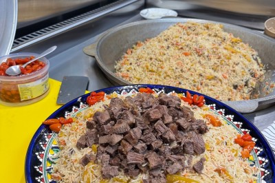 Un homme cuisine de grandes pièces de viande dans une marmite fumante en extérieur, portant un tablier et des gants.
