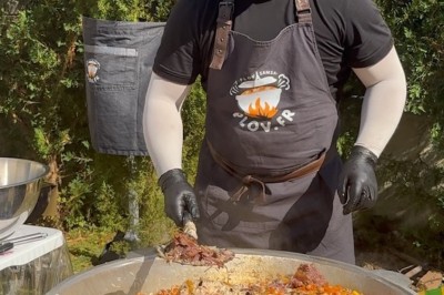Un homme cuisine de grandes pièces de viande dans une marmite fumante en extérieur, portant un tablier et des gants.
