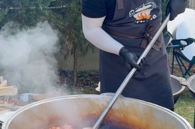 Un homme cuisine de grandes pièces de viande dans une marmite fumante en extérieur, portant un tablier et des gants.