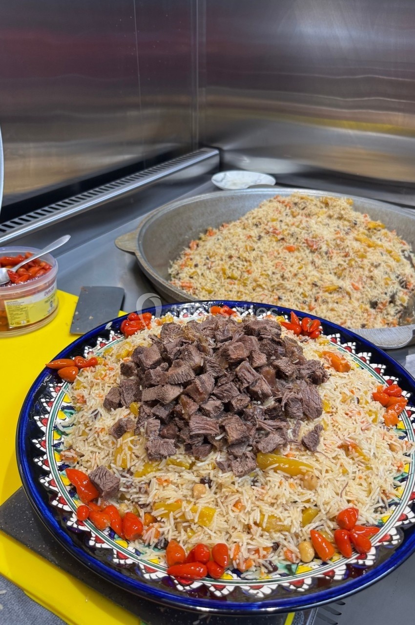 Plat de riz garni de morceaux de viande et de légumes, présenté dans une assiette décorée, avec une poêle en arrière-plan.