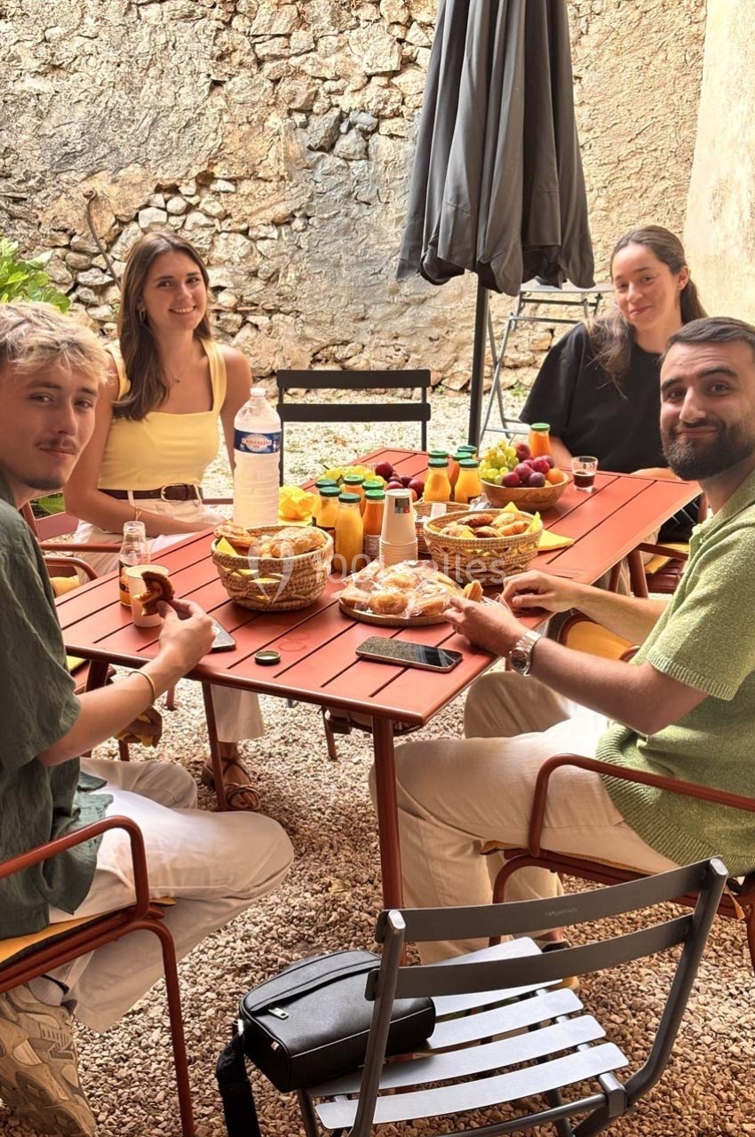 Quatre personnes assises autour d'une table en extérieur, partageant un petit-déjeuner avec fruits, jus et viennoiseries.