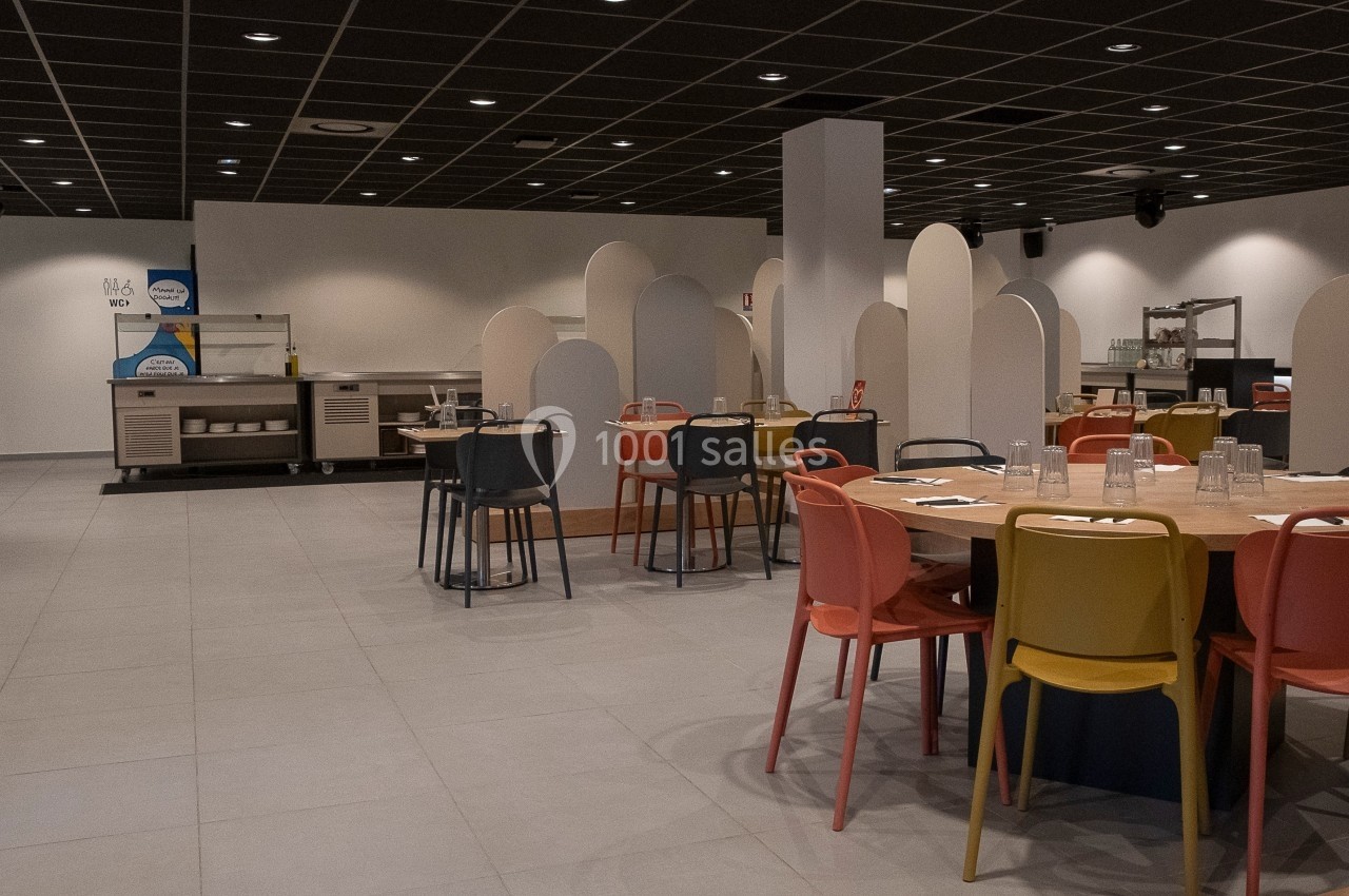Salle de restaurant moderne avec tables rondes, chaises colorées et cloisons de séparation sous un plafond noir.