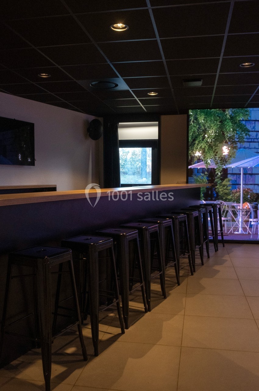 Bar intérieur avec un comptoir en bois, des tabourets hauts noirs et une vue sur une terrasse éclairée à l'extérieur.