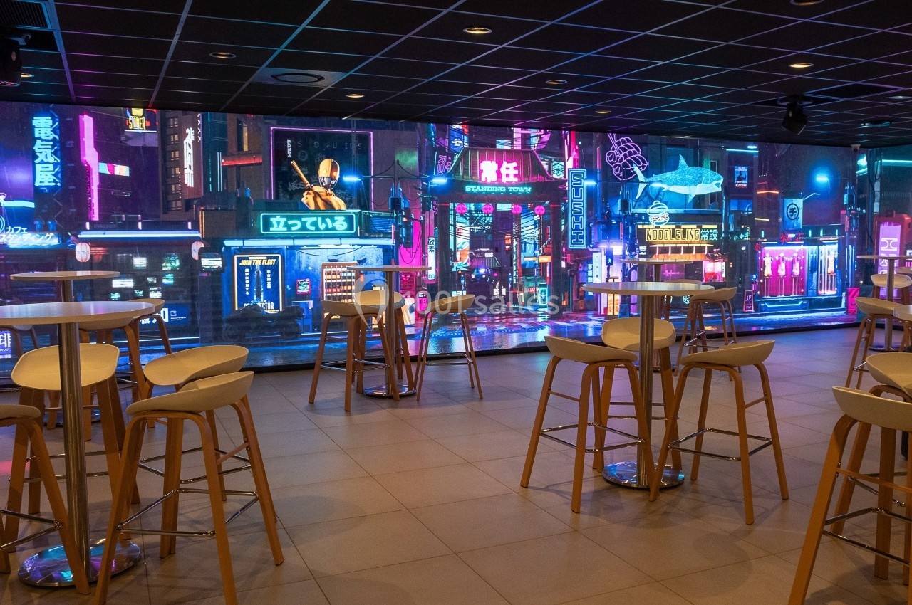 Salle moderne avec tables hautes et chaises, décorée d'une fresque lumineuse représentant une ville futuriste la nuit.