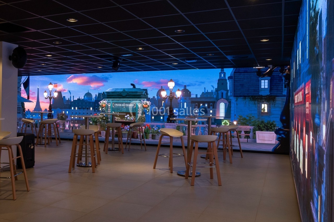 Salle moderne avec des tables hautes, des tabourets et un écran mural affichant une vue urbaine au coucher du soleil.