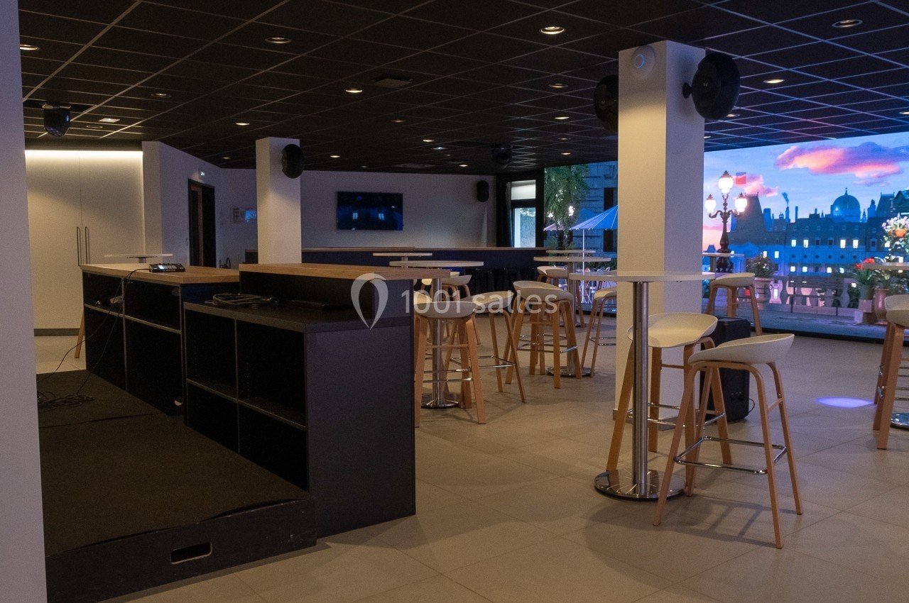 Salle moderne avec tables hautes, tabourets et grande baie vitrée donnant sur une vue urbaine éclairée.