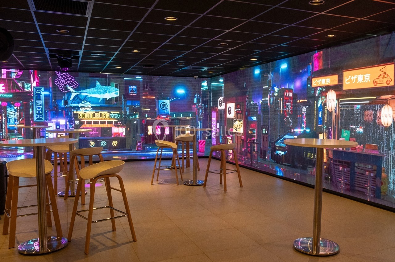 Salle moderne avec tables hautes et tabourets, décorée de panneaux lumineux inspirés d'une ambiance urbaine nocturne.