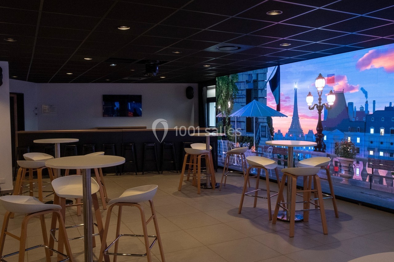 Salle moderne avec tables hautes, tabourets en bois et écran mural affichant une vue urbaine au coucher du soleil.