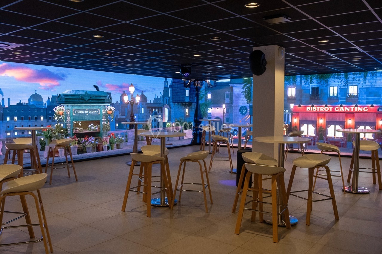 Salle moderne avec tables hautes et tabourets, vue sur une façade illuminée et un décor urbain en arrière-plan.