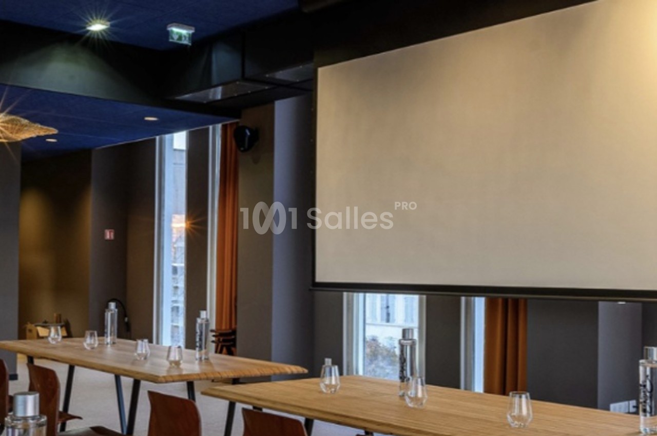 Salle de conférence avec tables en bois, écran de projection, murs gris clair et baies vitrées sur Marseille.