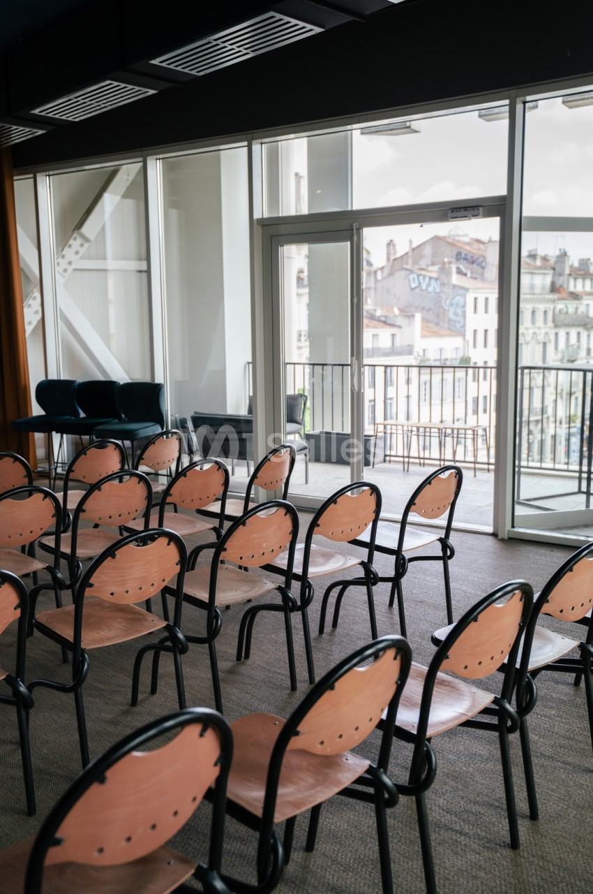 Salle avec chaises en bois et métal face à baies vitrées ouvertes sur terrasse et vue urbaine de Marseille.