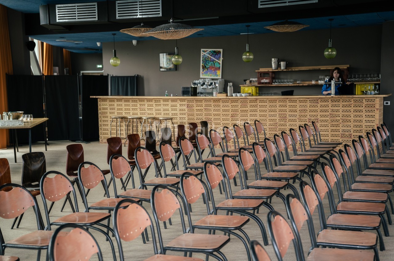 Salle intérieure avec bar en briques, chaises roses alignées face à un podium, lustre et éclairage suspendu, plafond bleu.
