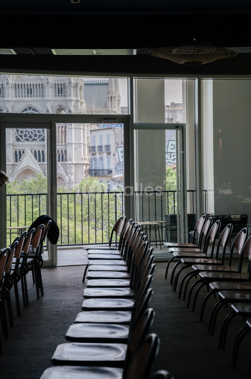 Salle vide avec chaises noires alignées face à une baie vitrée ouvrant sur une terrasse et la cathédrale de Marseille.