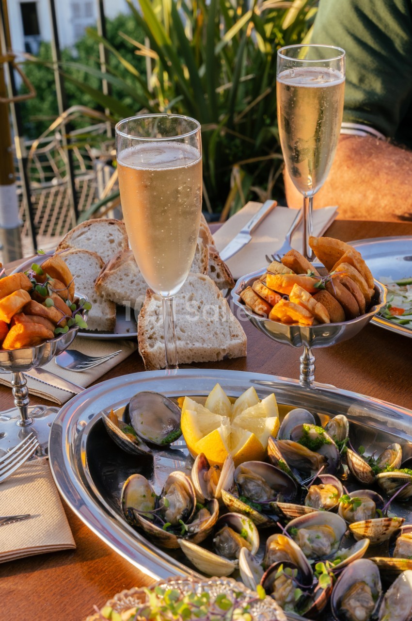 Table dressée en terrasse avec moules aux citrons, pain, verres de champagne et plantes - ciel bleu en arrière-plan.
