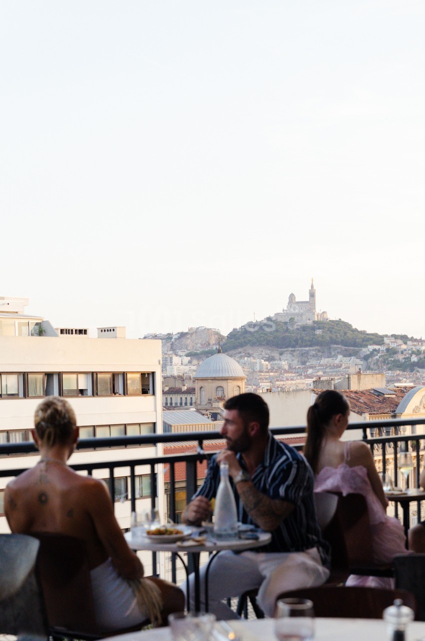 Terrasse avec vue sur Marseille, basilique Notre-Dame-de-la-Garde et convives attablés.