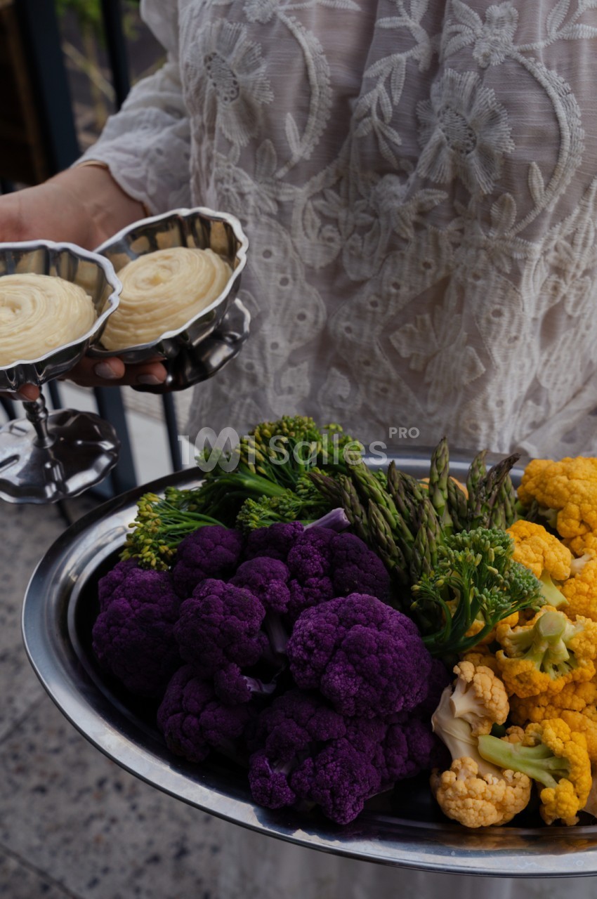 Personne en robe brodée tenant un plateau avec purée de pommes de terre et légumes colorés - brocoli, chou-fleur violet et orange.