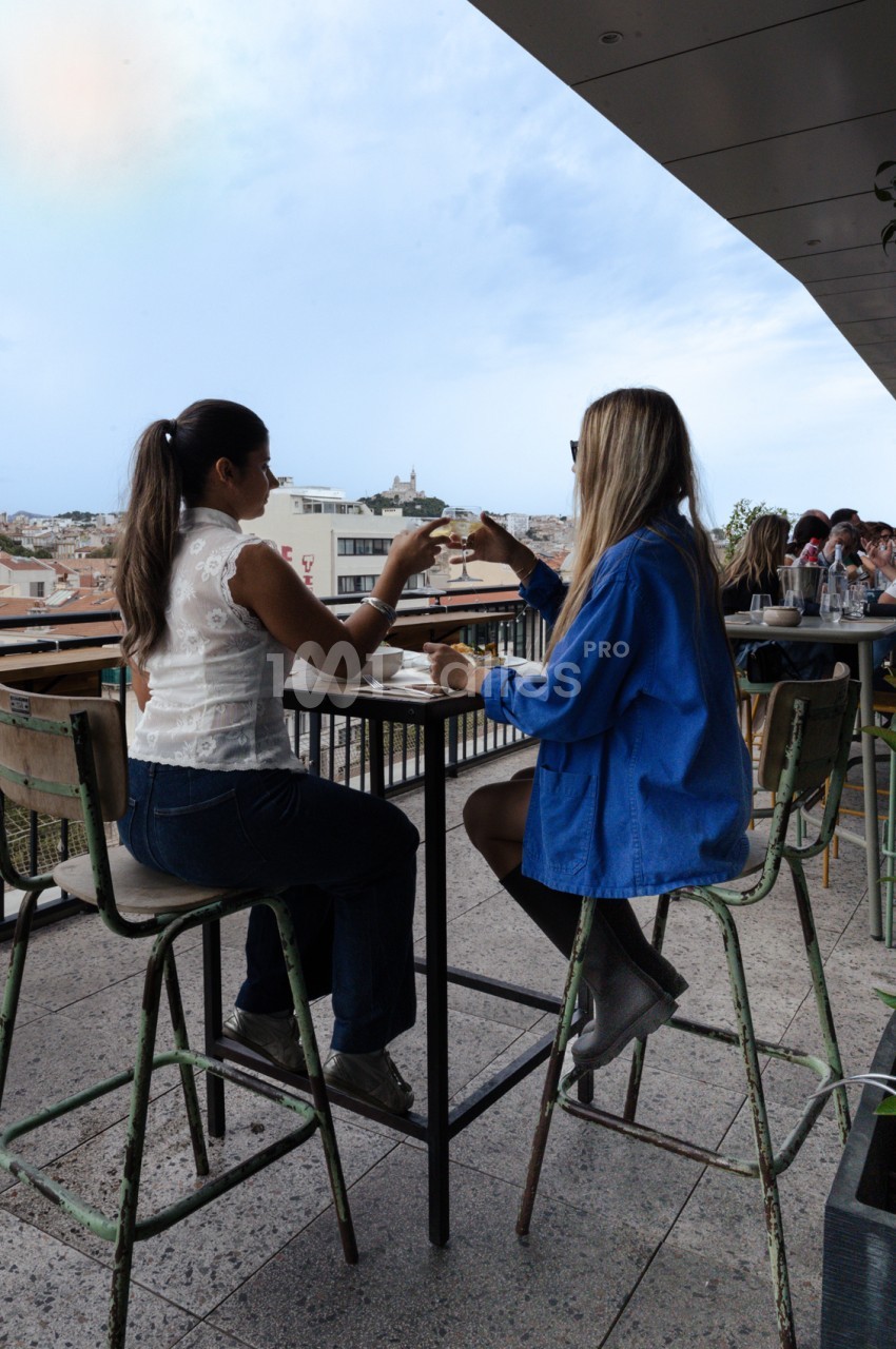 Terrasse en rooftop avec deux personnes assises à une table haute, vue panoramique sur Marseille et la basilique Notre-Dame.