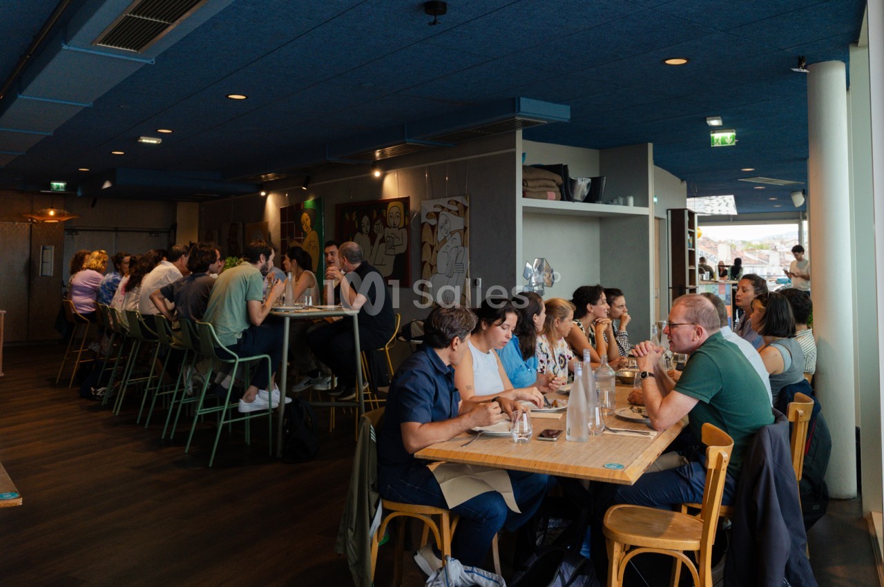 Salle intérieure avec plafond bleu, tables en bois et chaises colorées, environ 50 convives en repas ou pause.