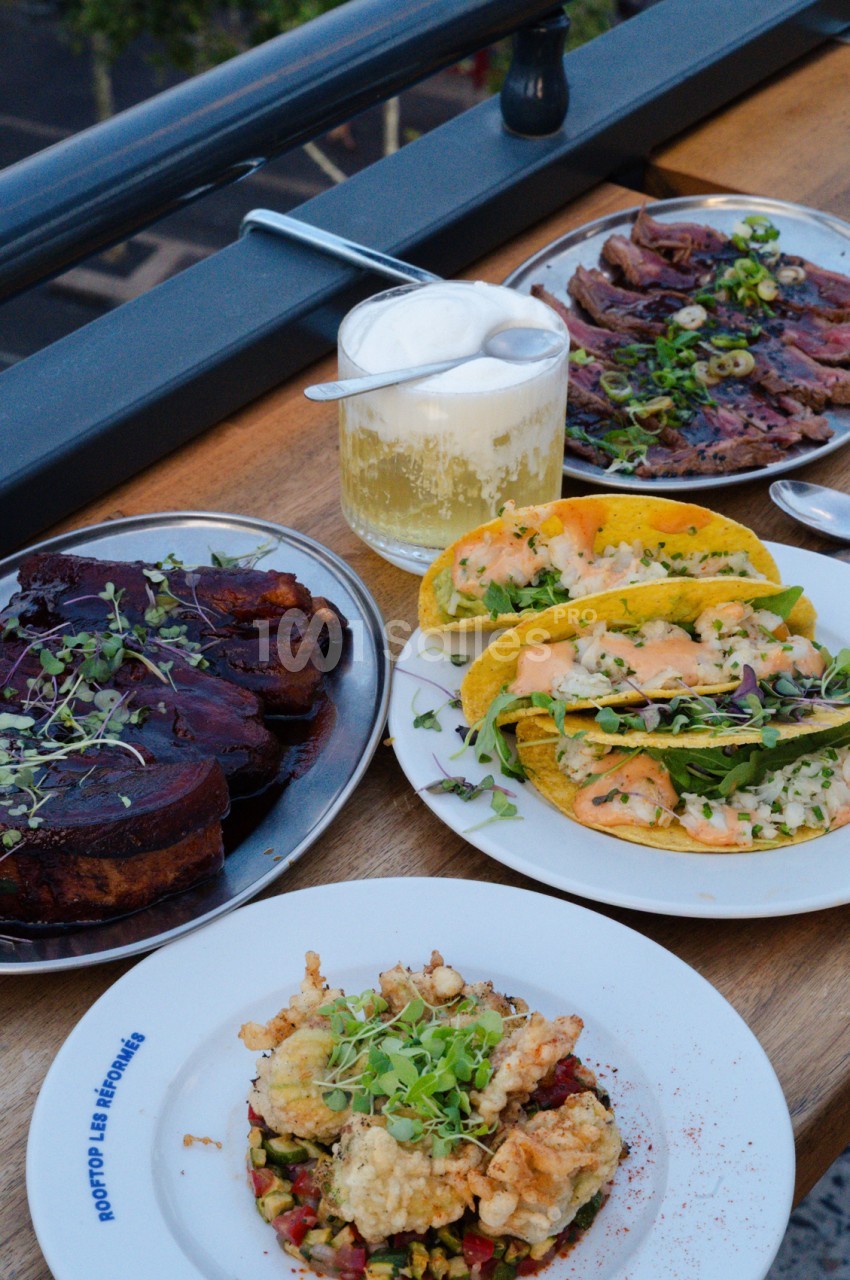 Assiettes de viandes et tacos garnis sur table en bois avec verre de vin blanc et vue sur la ville.