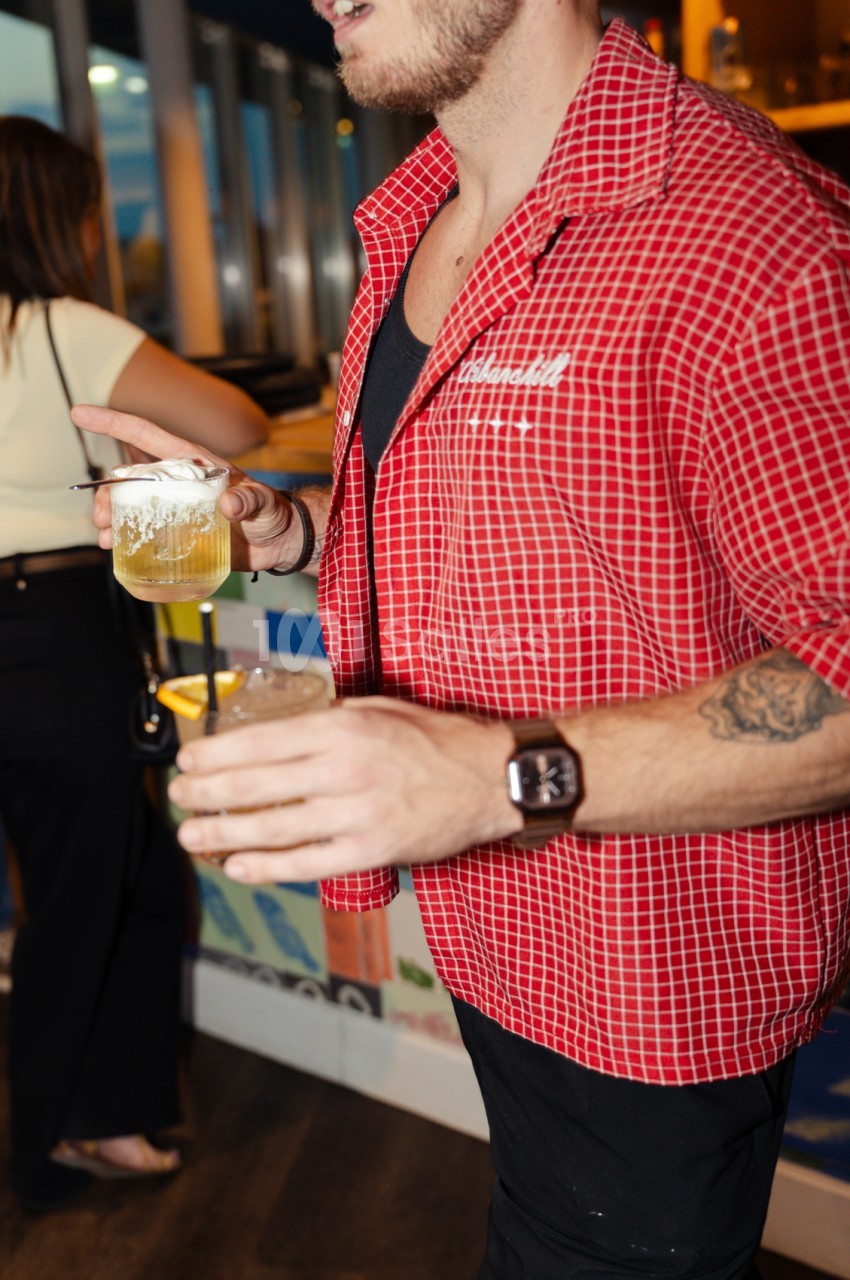 Homme en chemise à carreaux rouge tenant un verre de cocktail dans un espace intérieur chaleureux.