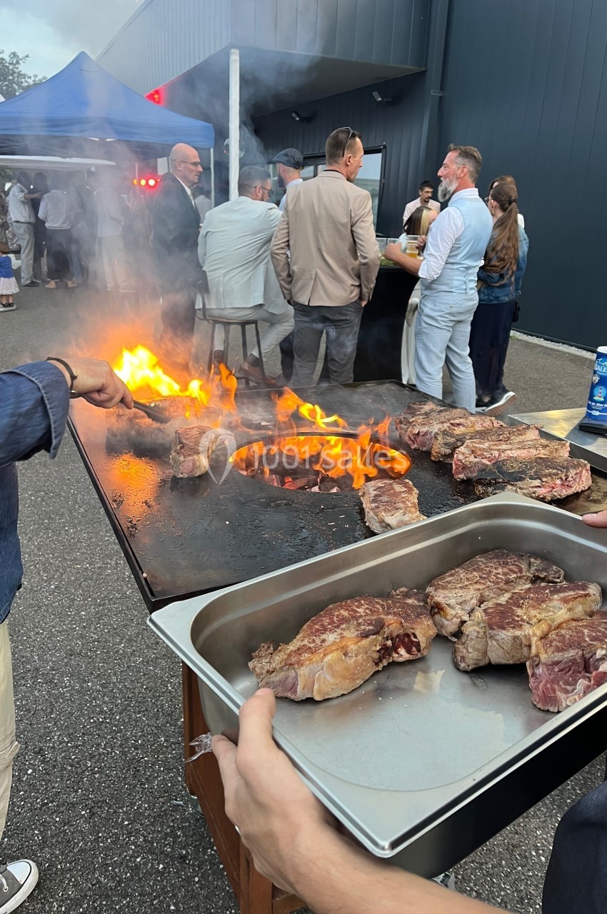 Personnes rassemblées autour d'un barbecue en plein air, avec des steaks en cuisson sur une grande plaque chauffante.