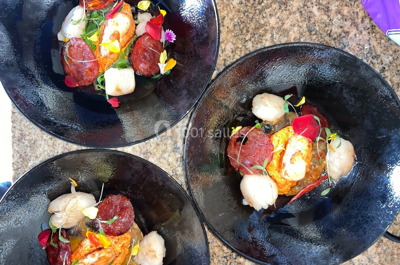 Trois assiettes noires contenant des pétoncles, tranches de chorizo, légumes et fleurs comestibles sur une table en granit.