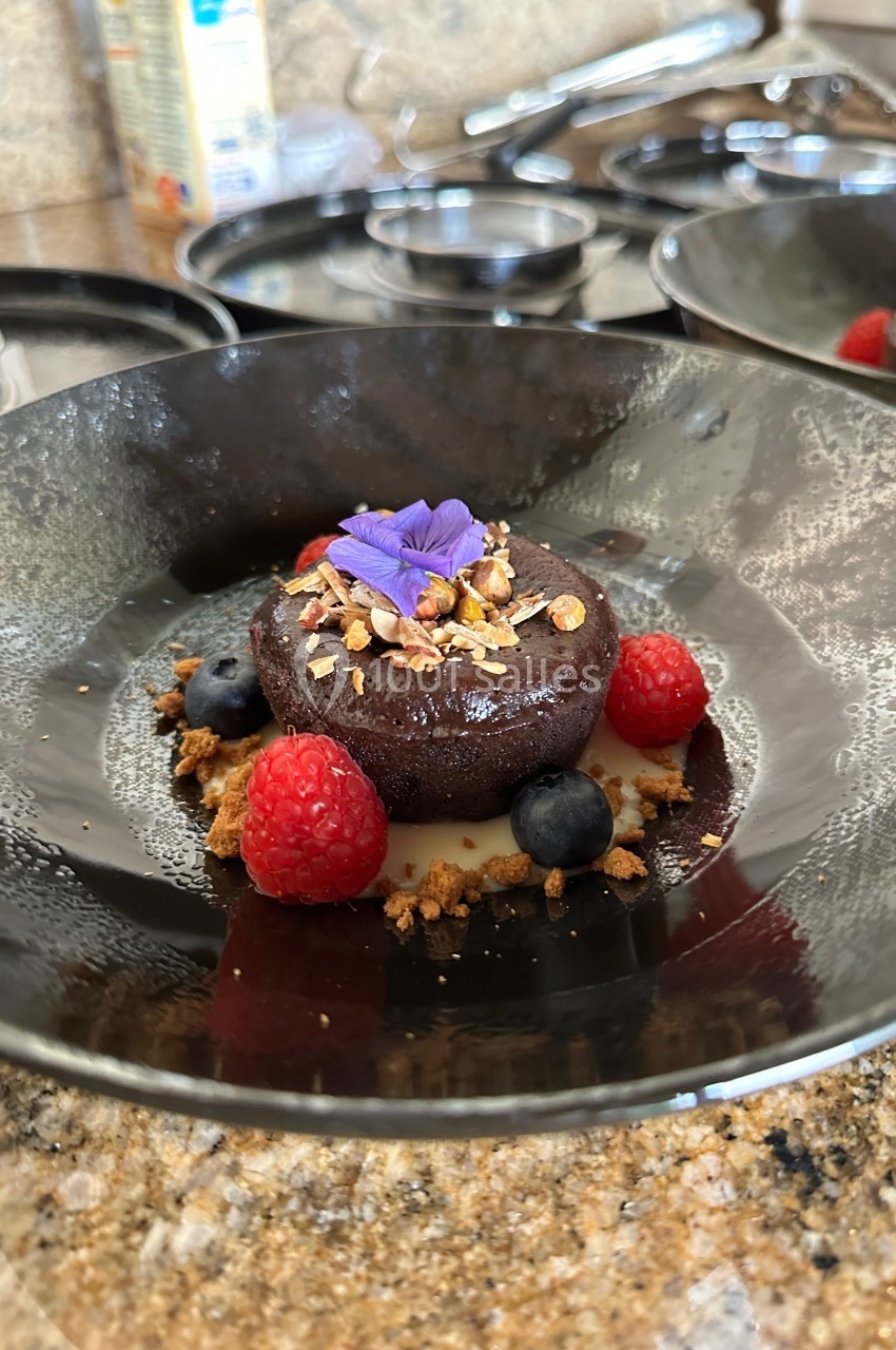 Dessert au chocolat décoré de fruits rouges, myrtilles, granola et une fleur comestible, servi dans une assiette noire.