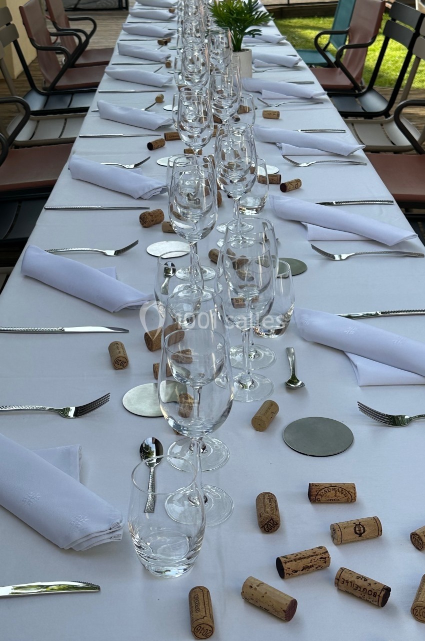 Table dressée avec nappes blanches, verres à vin, couverts, serviettes pliées et bouchons de liège dispersés.