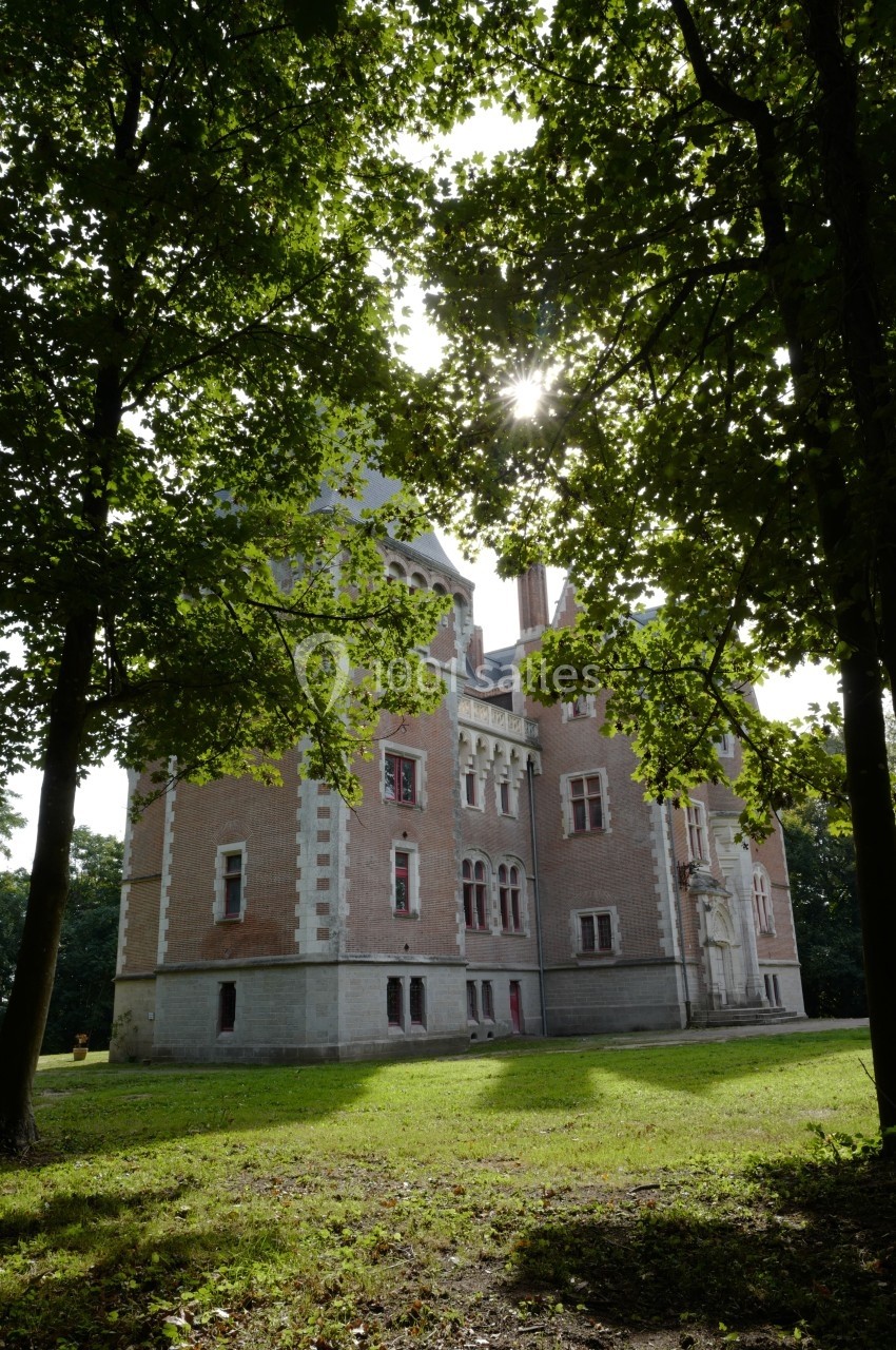 Château en briques entouré d'arbres, avec des rayons de soleil traversant le feuillage.