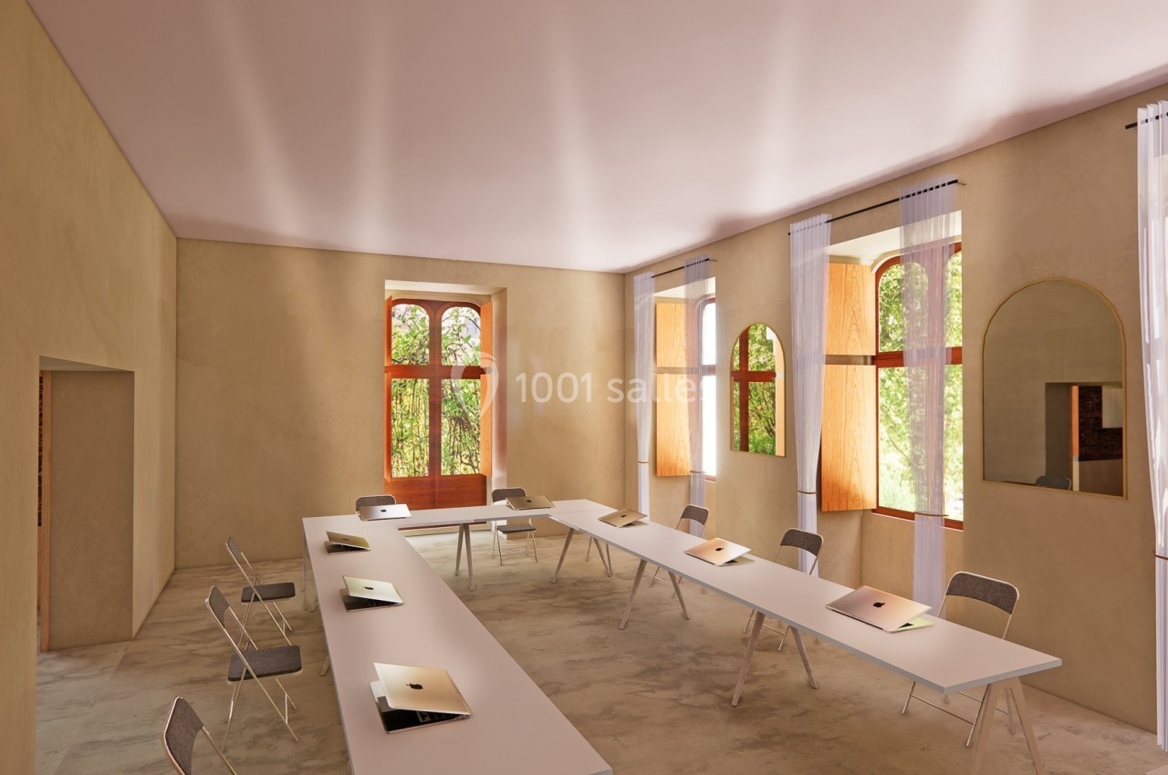 Salle lumineuse avec de grandes fenêtres, tables blanches alignées, chaises pliantes et ordinateurs portables posés.