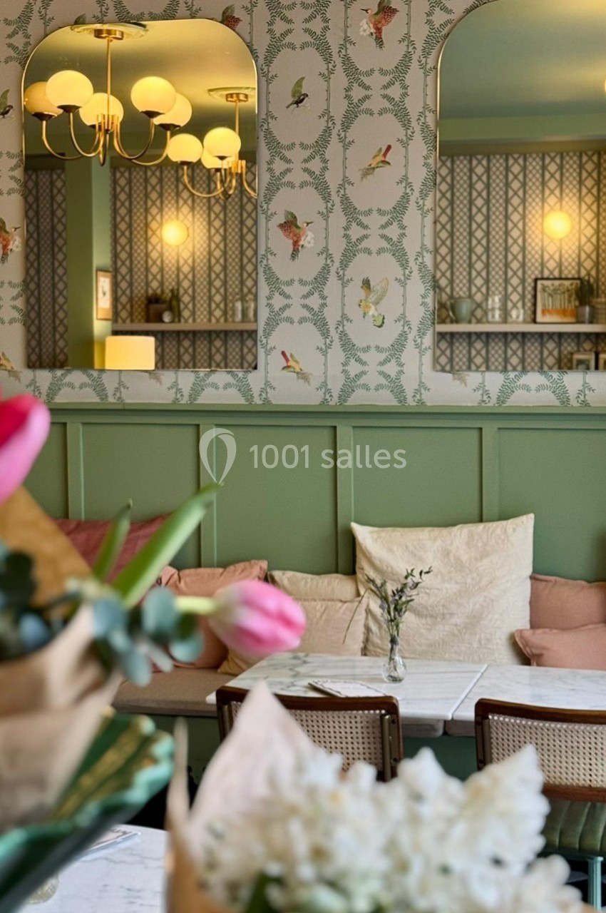 Intérieur d'un café avec banquettes vertes, coussins pastel, fleurs en premier plan et papier peint à motifs d'oiseaux.
