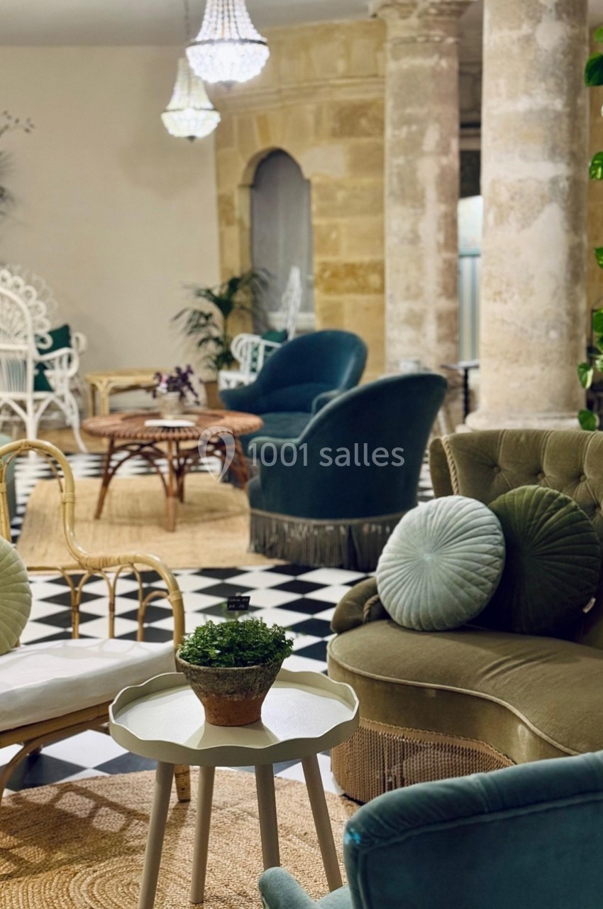 Salon élégant avec fauteuils en velours, table basse, plantes et sol en damier noir et blanc, éclairé par des lustres.