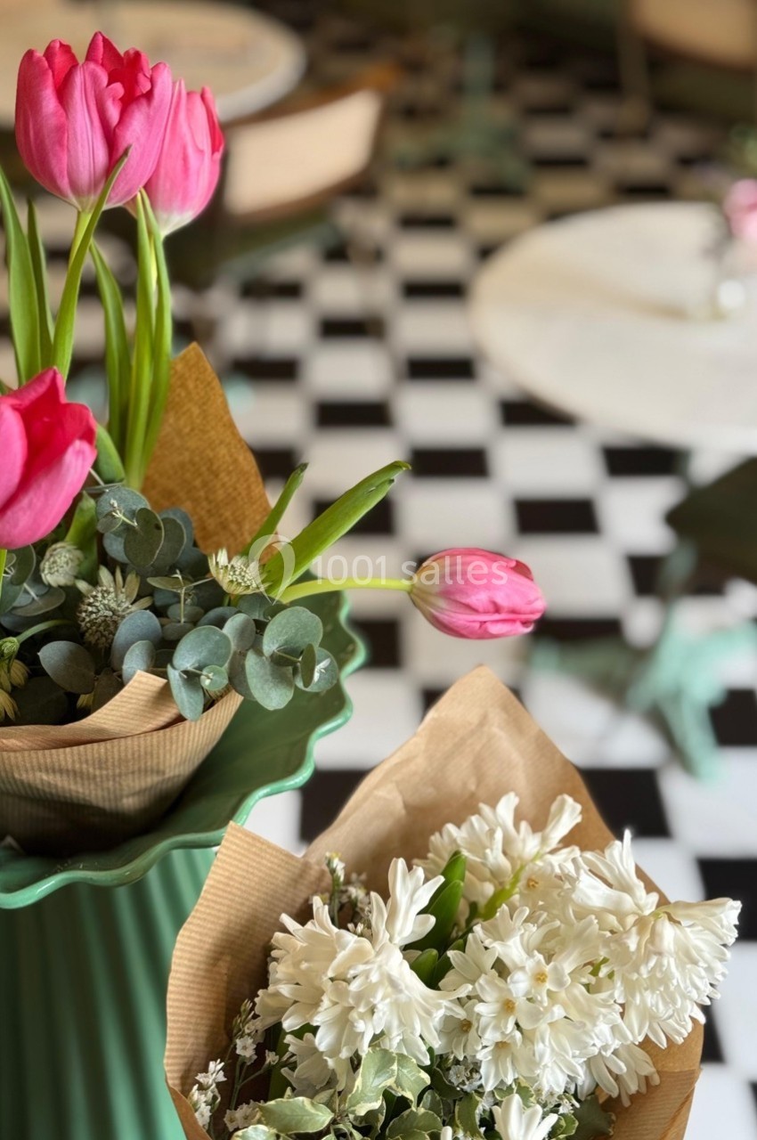 Bouquets de tulipes roses et de fleurs blanches sur un sol en damier noir et blanc, avec des tables floues en arrière-plan.