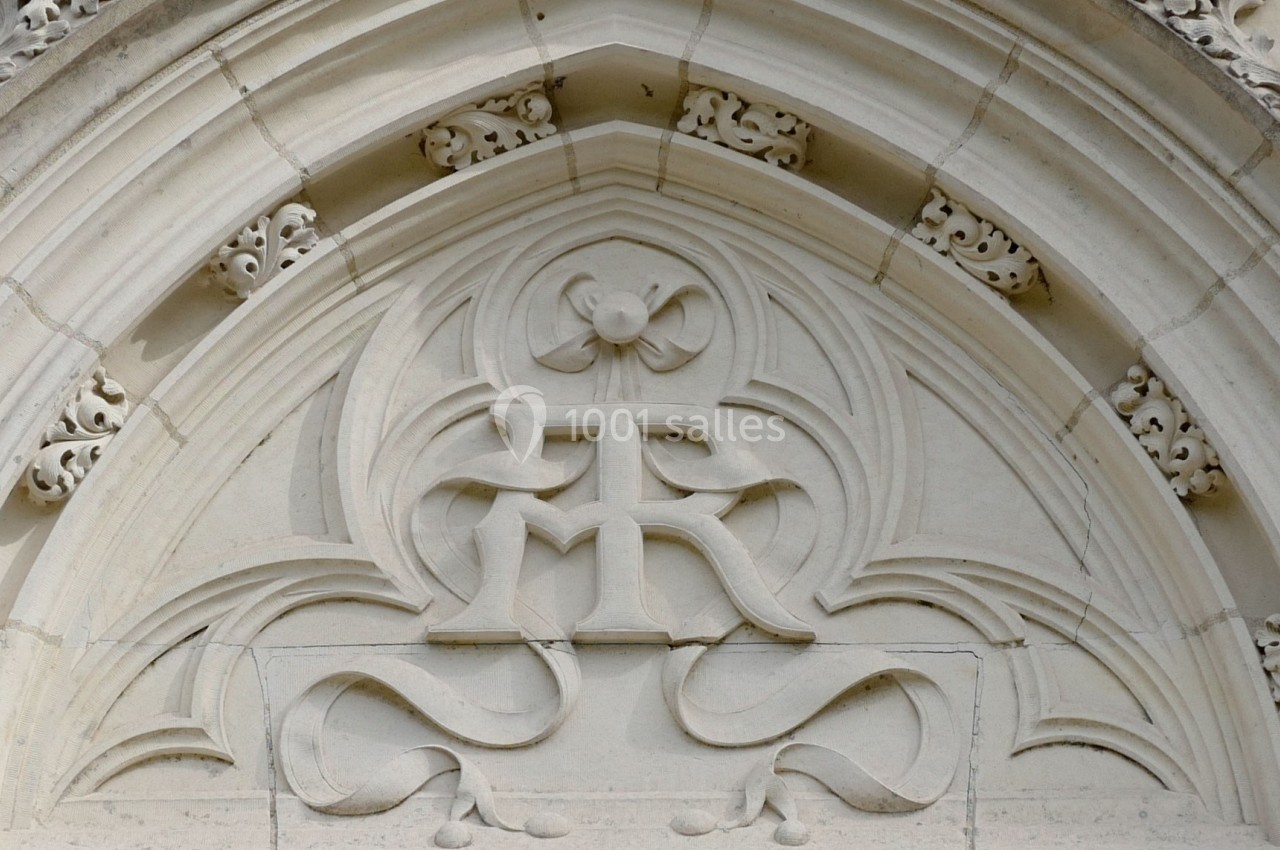 Bas-relief sculpté en pierre représentant un monogramme orné de motifs floraux dans une arche gothique.