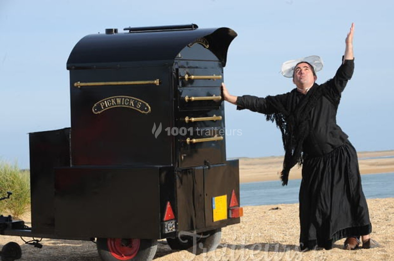 Un homme en costume traditionnel pose de manière théâtrale près d'un four mobile noir sur une plage.