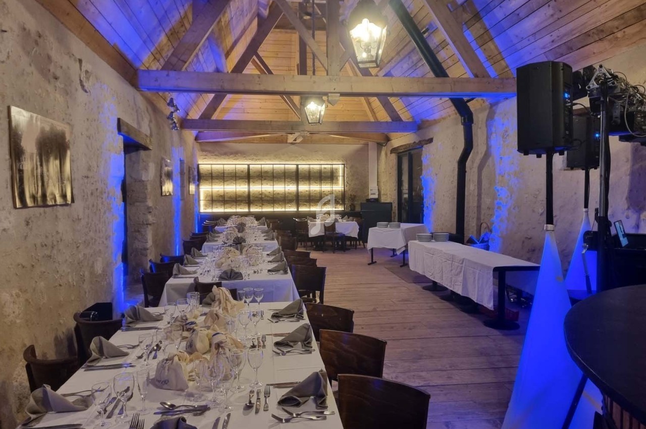 Salle de réception en pierre avec poutres apparentes, tables dressées pour un repas, éclairage bleu et chaleureux.