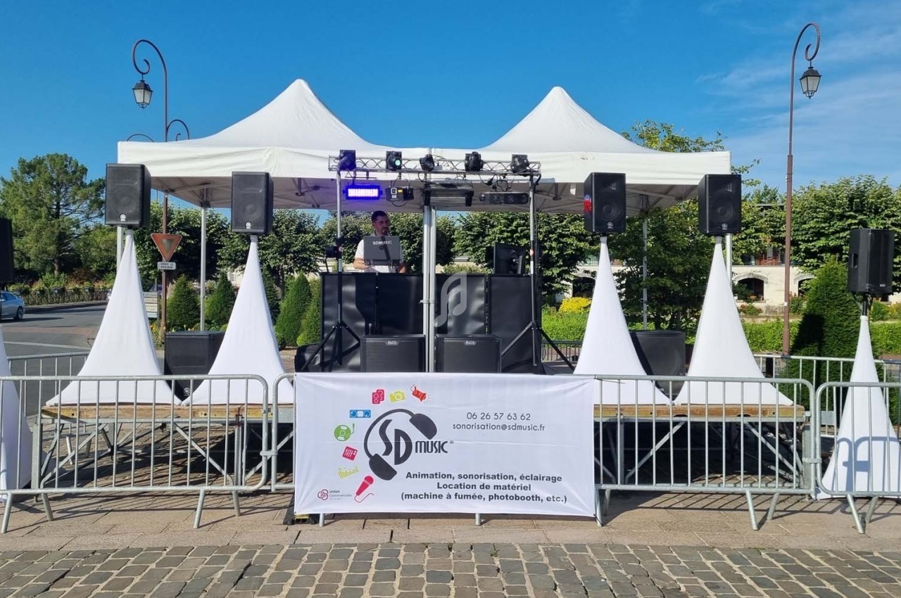 Scène extérieure avec un DJ sous des tentes blanches, entourée de barrières et équipée de matériel d'éclairage.