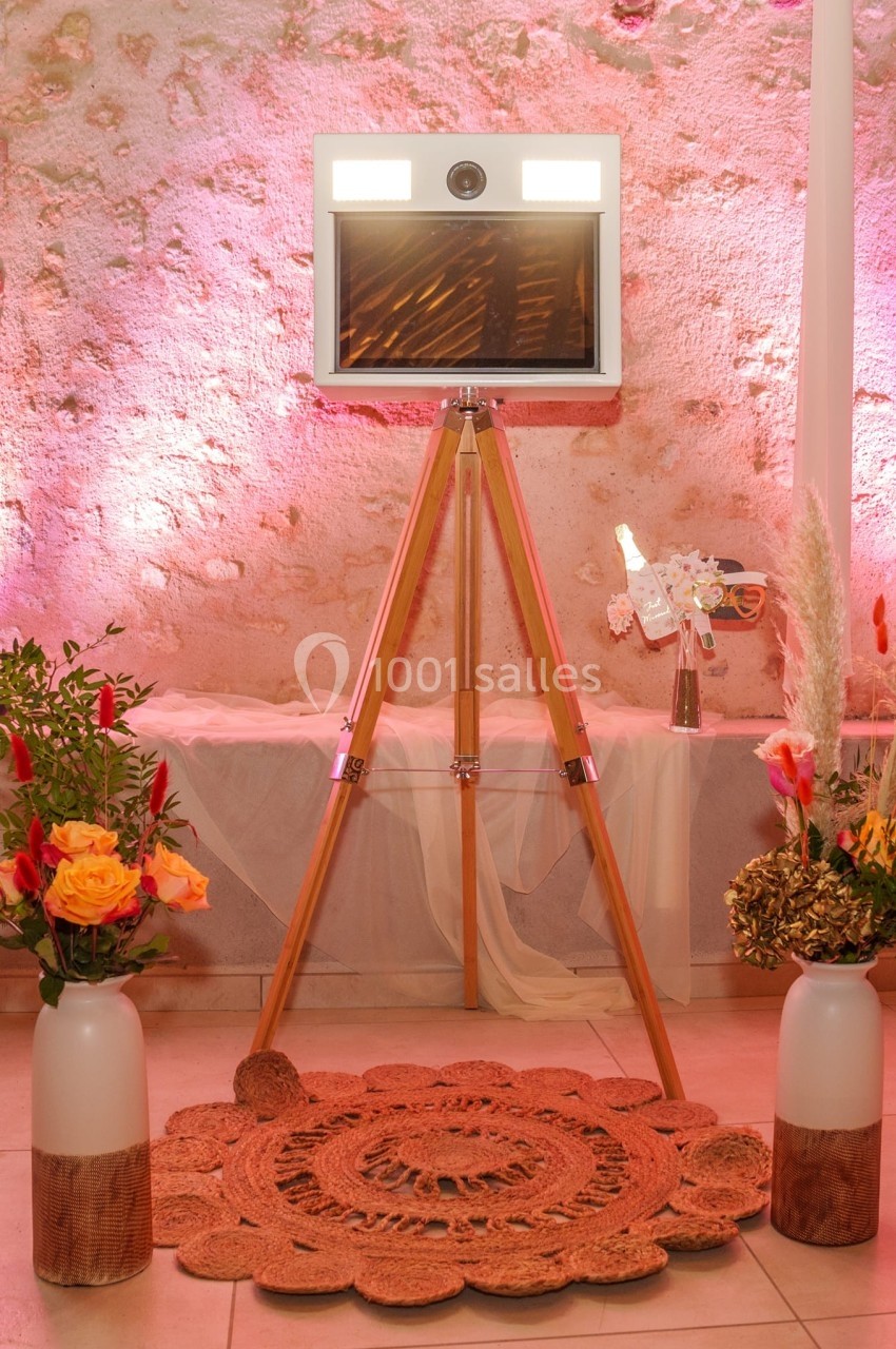 Un photobooth sur trépied en bois entouré de décorations florales et éclairé par des lumières roses.