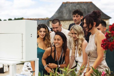 Borne photo blanche sur pied avec écran et imprimante intégrée, installée en extérieur sur une terrasse ensoleillée.