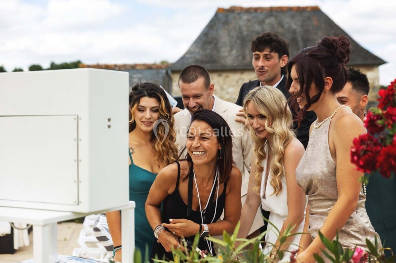 Un groupe de personnes souriantes regarde un écran lors d'un événement en extérieur, près de fleurs rouges.