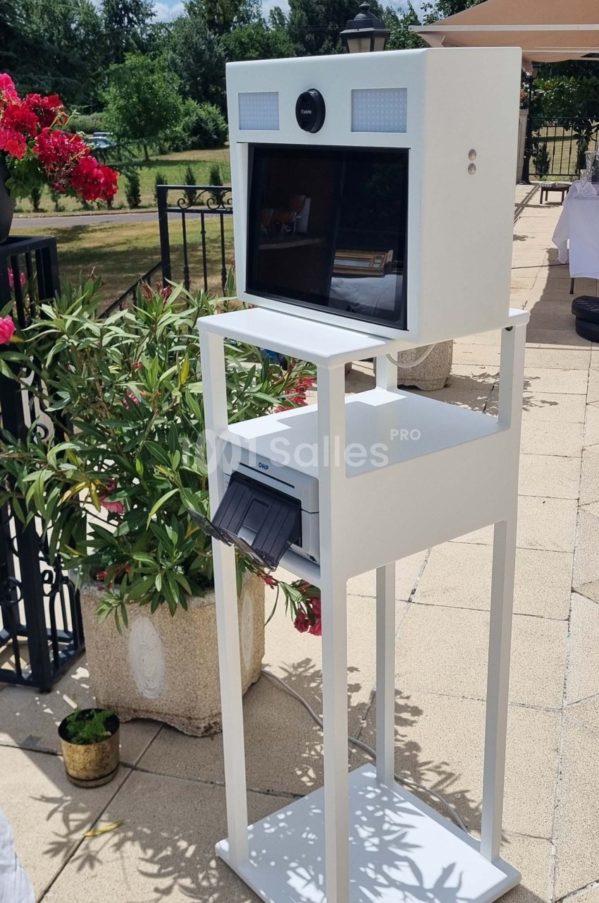 Borne photo blanche sur pied avec écran et imprimante intégrée, installée en extérieur sur une terrasse ensoleillée.