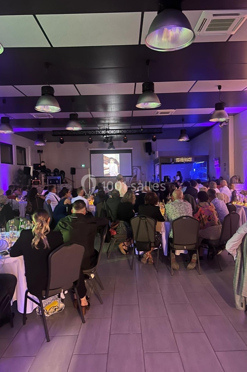 Salle de réception avec des invités assis à des tables rondes, regardant une projection sur écran.